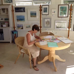 Sara writing at a round table with her artwork displayed in the background