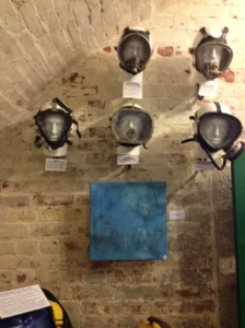Sara's art displayed on a brick wall at the underwater art exhibition. Above the artwork, 5 diving helmets are displayed on dummy heads.
